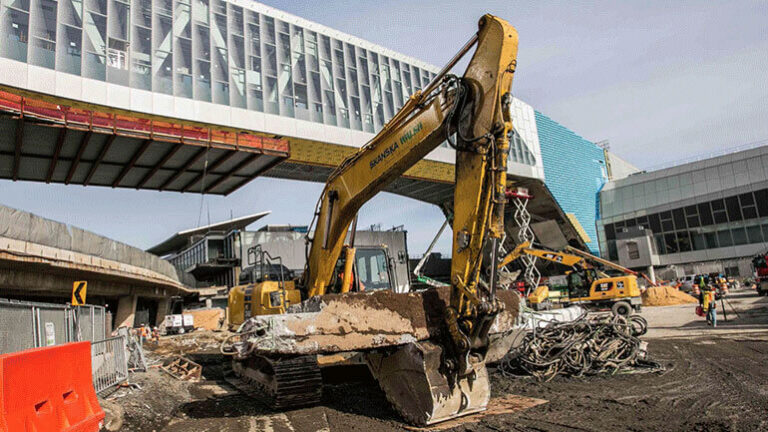 An excavator sits in front of a Terminal B pedestrian bridge